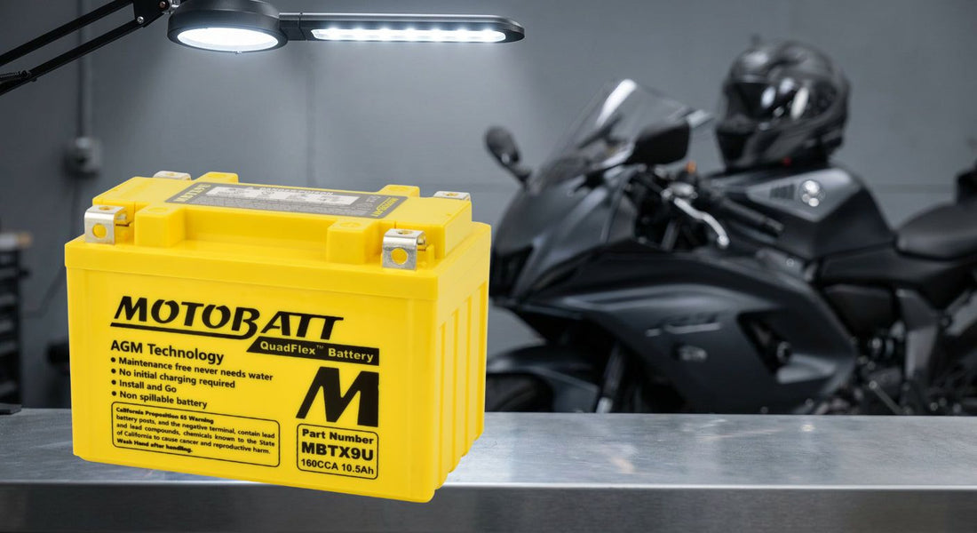 A black AGM powersport battery on a workshop bench with a motorcycle visible in the background.