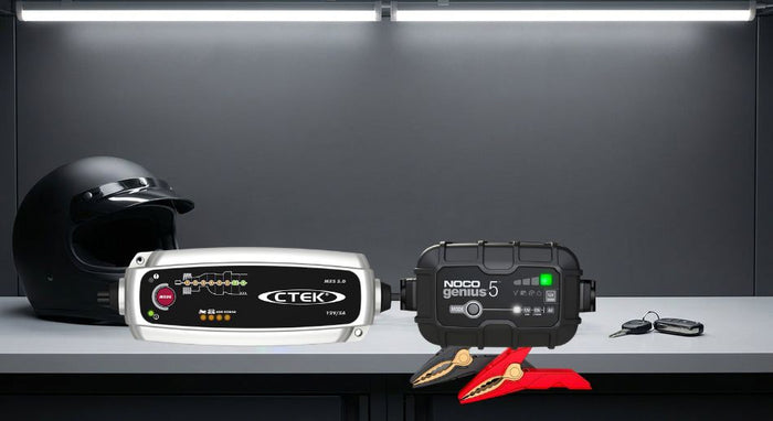 Two smart battery chargers placed side by side on a workshop bench alongside a motorcycle helmet and car keys, lit with clean studio lighting against a dark grey background.