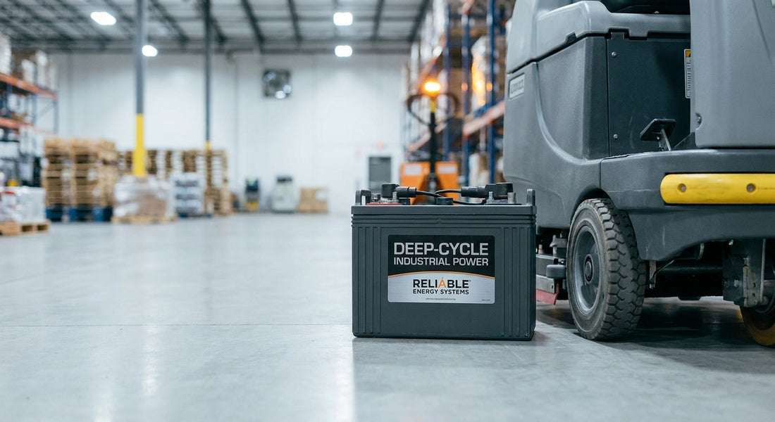A deep-cycle traction battery positioned beside the wheel of an industrial floor scrubber on a polished concrete warehouse floor.