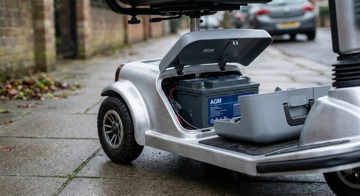 A mobility scooter parked outdoors with its battery compartment open, revealing a clean AGM battery inside, photographed in soft natural daylight.