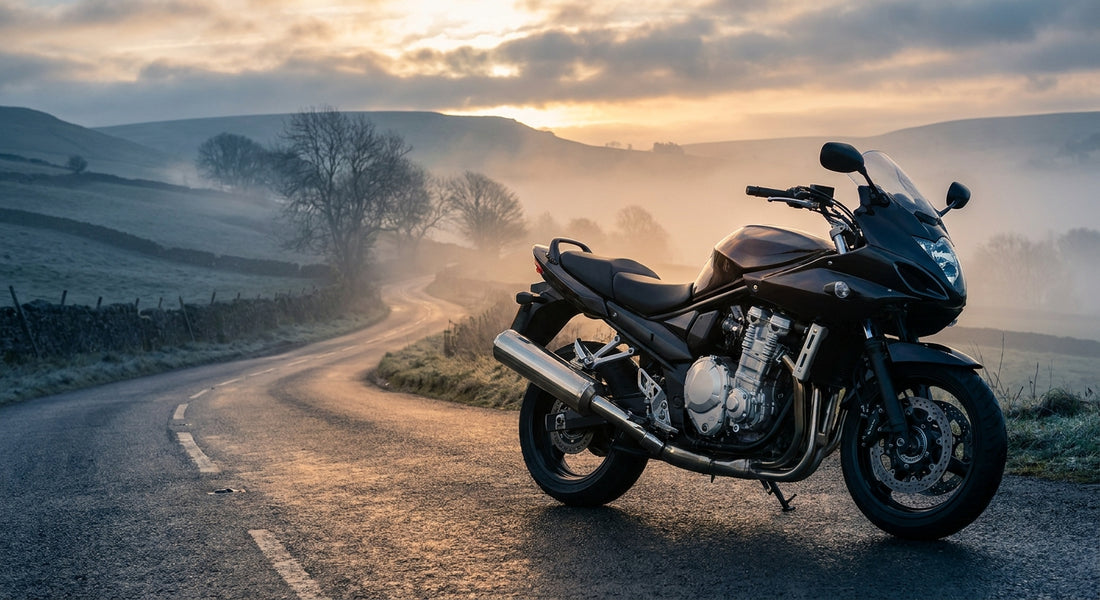 A modern motorcycle parked on a misty British country road at dawn, bathed in soft golden and cool blue-grey light.